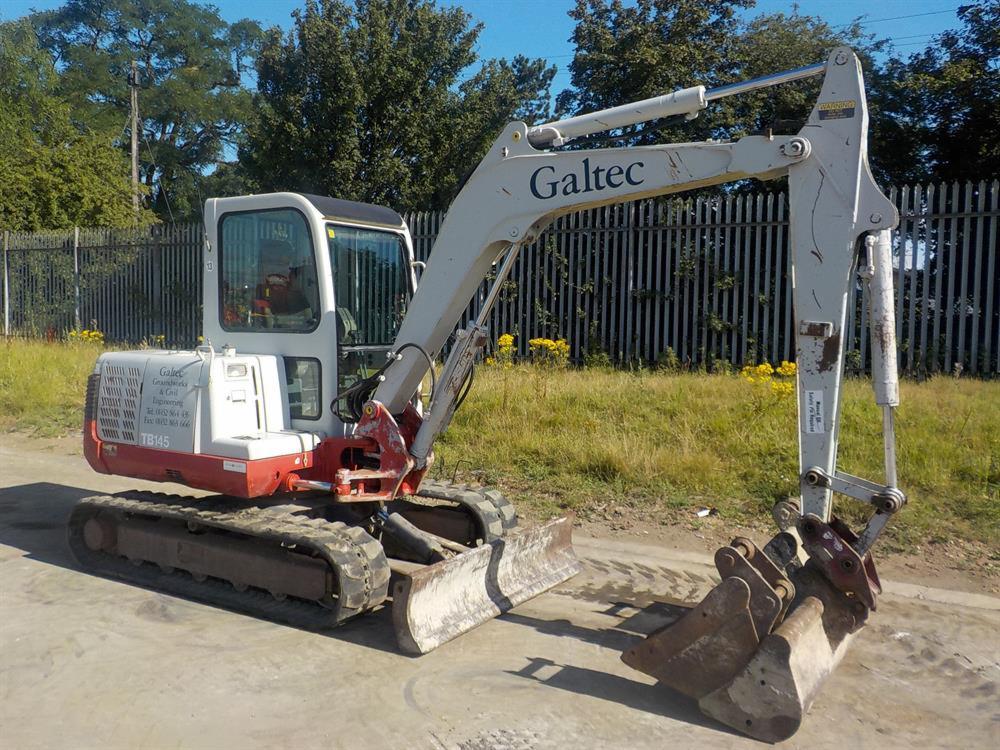 Kompaktowa koparka Takeuchi TB145 z łyżką wielofunkcyjną, stojąca na utwardzonym podłożu, z widocznym logo Galtec na ramieniu, na tle metalowego ogrodzenia i zieleni.