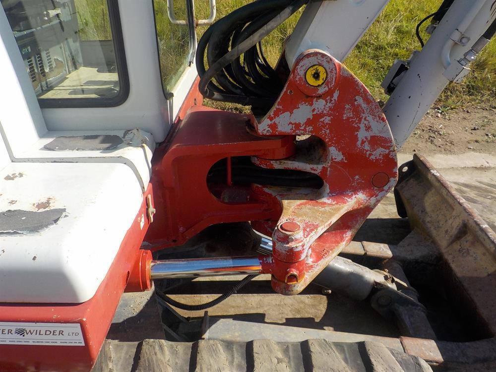 Zbliżenie na ramię robocze minikoparki Takeuchi TB145 z widocznymi śladami użytkowania, farba w kolorze czerwonym i szarym odpryskuje, na pierwszym planie fragment kabiny i gąsienicy.