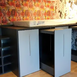 Wielofunkcyjna konstrukcja meblowa w pokoju młodzieżowym: łóżko na antresoli z materacem w folii, schody z półkami, szafy z szarymi frontami i otwartą przestrzenią pod łóżkiem, tapeta z motywem...