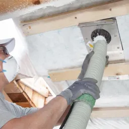 Pracownik w masce i rękawicach aplikuje izolację za pomocą węża do otworu w dachu, widoczne elementy drewnianej konstrukcji i folia paroizolacyjna.