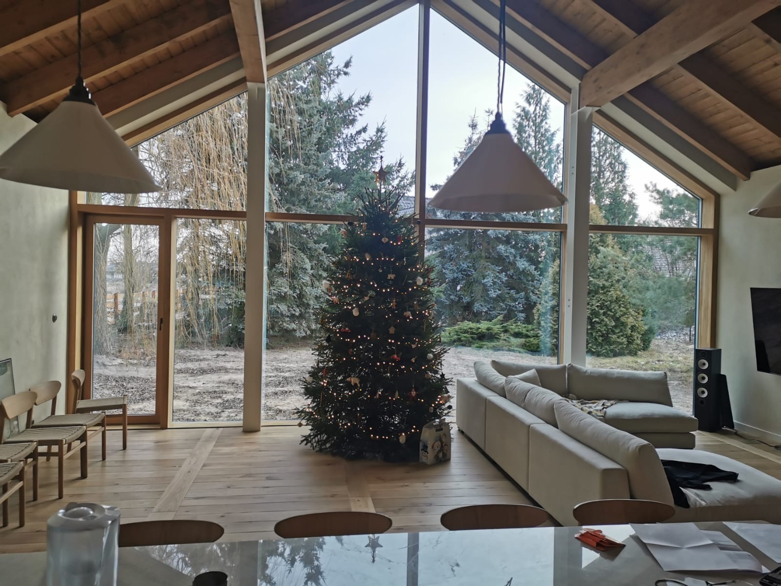 Jasne wnętrze domu z drewnianą konstrukcją dachu, dużymi oknami z widokiem na zimowy ogród i choinką w rogu, oświetlone wiszącymi lampami.