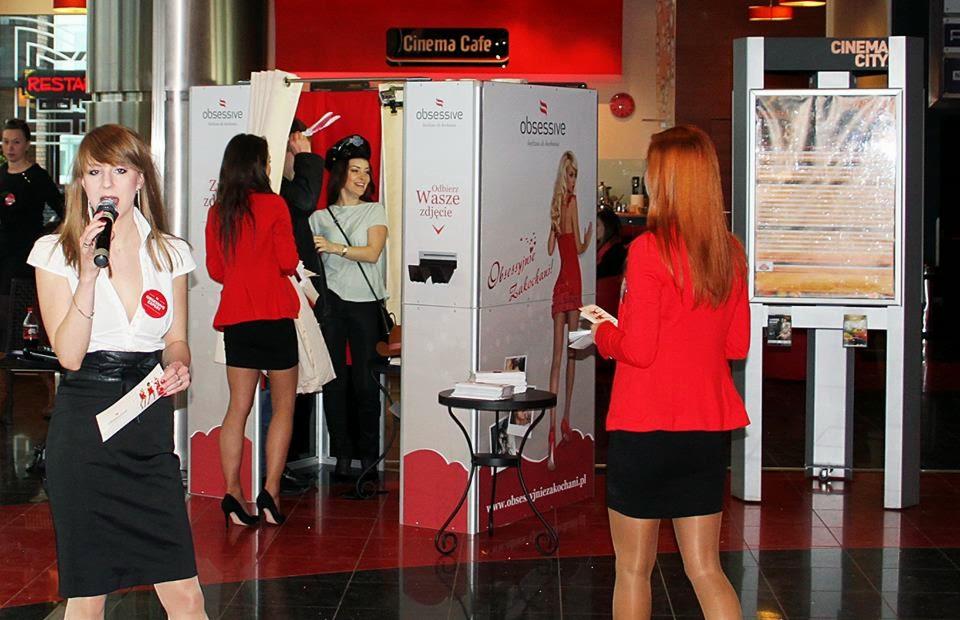 Wnętrze galerii handlowej z fotobudką marki Obsessive; prezenterka z mikrofonem w dłoni, w tle osoby korzystające z fotobudki i automat z ramkami do zdjęć.