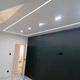 Nowoczesne wnętrze z podświetlanym sufitem i ciemną ścianą, widoczne otwarte drzwi do kolejnego pomieszczenia z drabiną w tle.