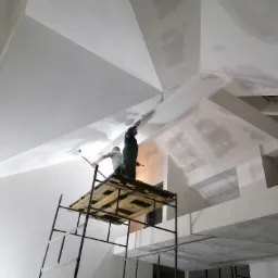 Malowanie skosów sufitu na wysokości z rusztowania przez dwóch malarzy, widoczne niedomalowane fragmenty i przewody oświetleniowe.