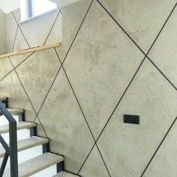 Nord-Bud - Ściana w korytarzu wykończona gładzią dekoracyjną w odcieniach szarości, z czarnymi liniami tworzącymi geometryczny wzór rombów, widoczne schody zabezpieczone folią ochronną.