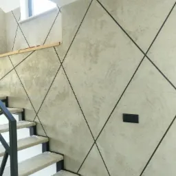 Ściana w korytarzu wykończona gładzią dekoracyjną w odcieniach szarości, z czarnymi liniami tworzącymi geometryczny wzór rombów, widoczne schody zabezpieczone folią ochronną.