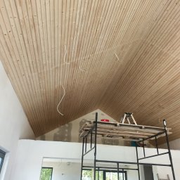 Nord-Bud - Drewniana boazeria na skośnym suficie poddasza, widoczne kable elektryczne, rusztowanie z palet i drabina w tle, ślady szpachlowania na ścianie.
