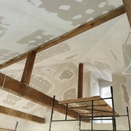 Nord-Bud - Skośny sufit z drewnianymi belkami w trakcie szpachlowania, widoczny fragment rusztowania.