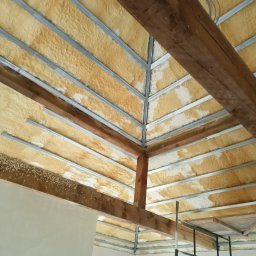 Nord-Bud - Poddasze z widoczną konstrukcją metalową i drewnianą, pokryte żółtą pianką izolacyjną z białymi plamami, widok od dołu.