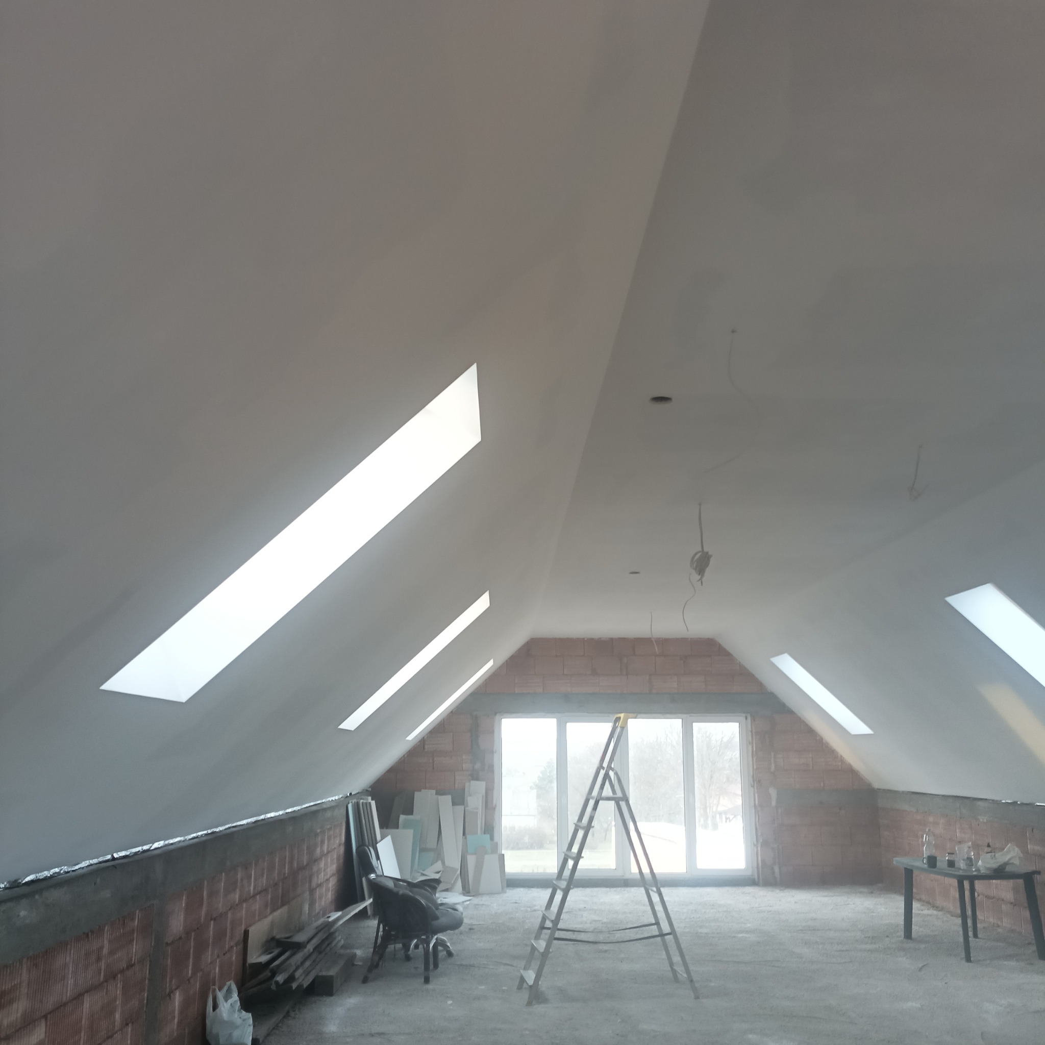 Poddasze w trakcie wykańczania: ściany z surowej cegły, częściowo pokryte gładzią, okna dachowe, drabina aluminiowa, widoczne przewody elektryczne, przygotowane płyty gipsowe.