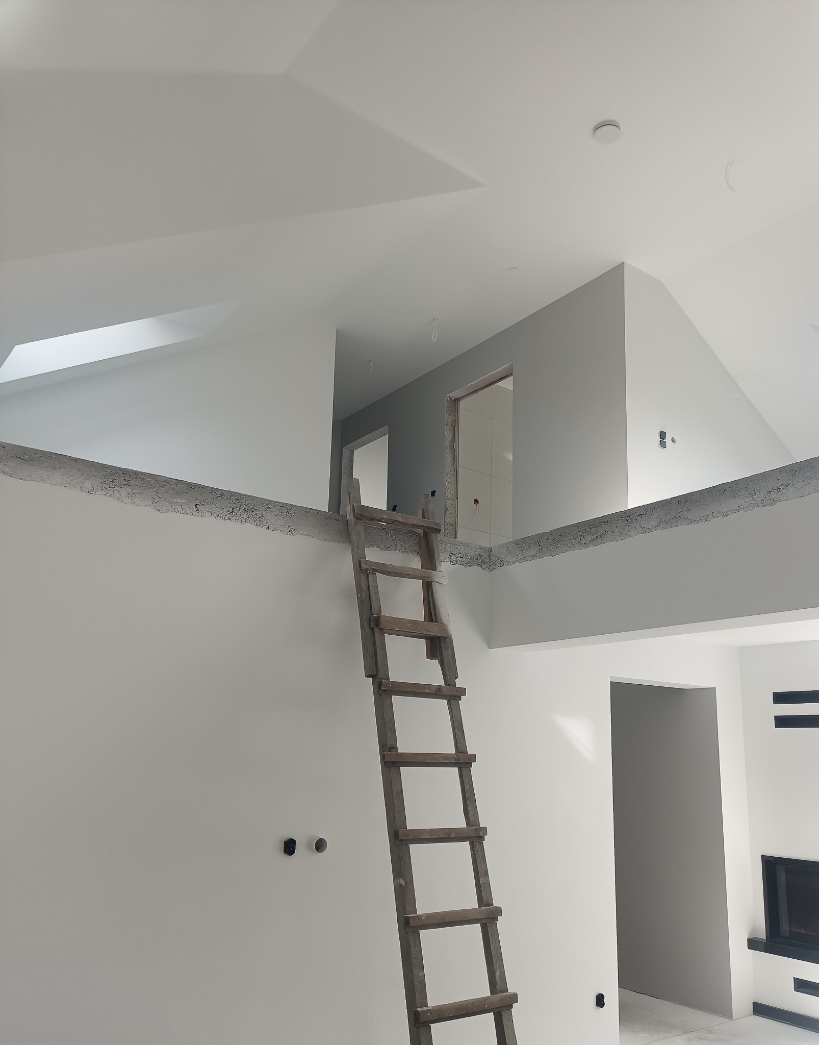 Wnętrze domu w trakcie wykańczania, z drewnianą drabiną opartą o ścianę, widoczne gładkie, świeżo pomalowane ściany i sufit, oraz otwory drzwiowe prowadzące do innych pomieszczeń.