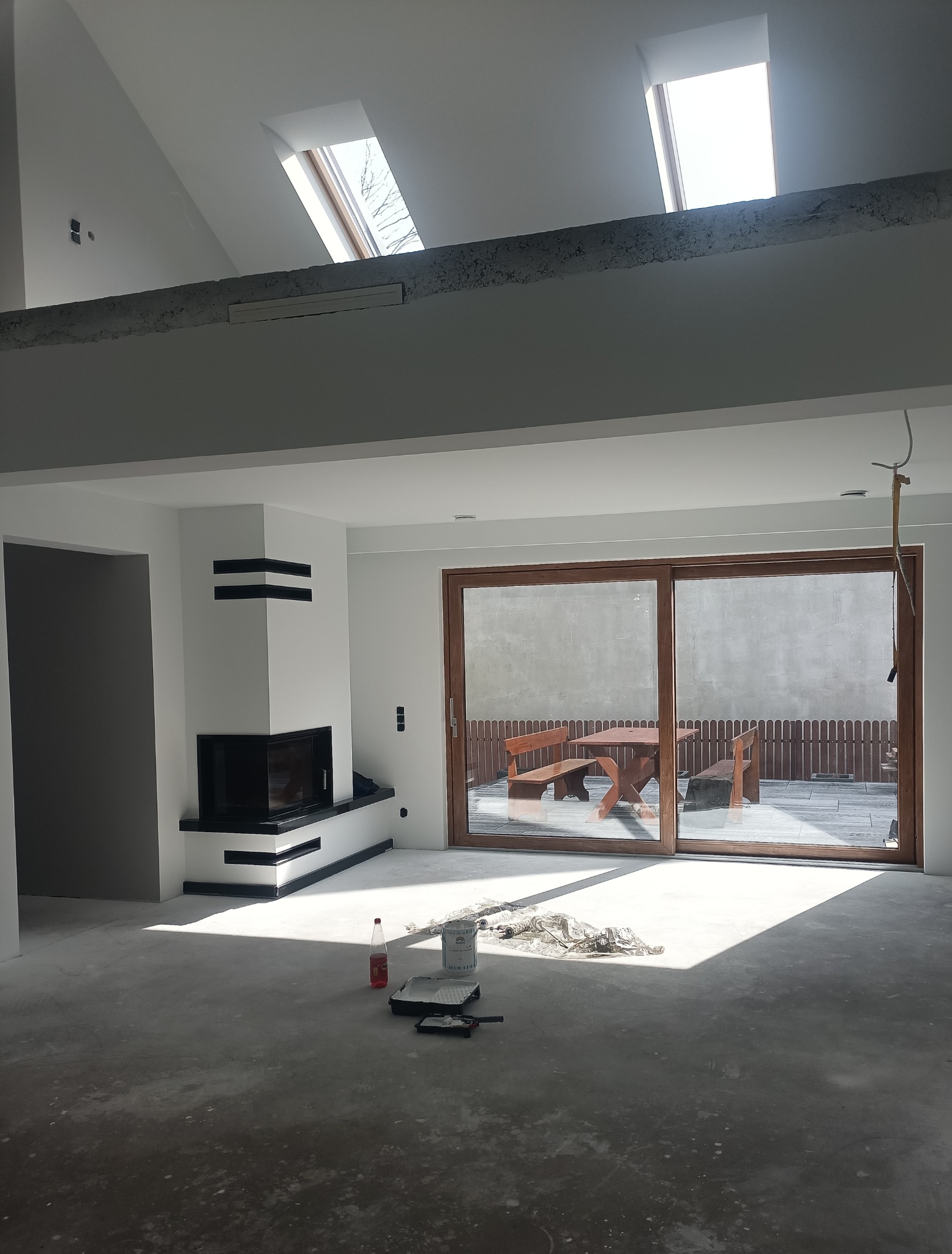 Wnętrze w trakcie wykańczania z widocznym kominkiem z czarnymi akcentami, oknami dachowymi i rozsuwanymi drzwiami na taras, z narzędziami malarskimi na podłodze.