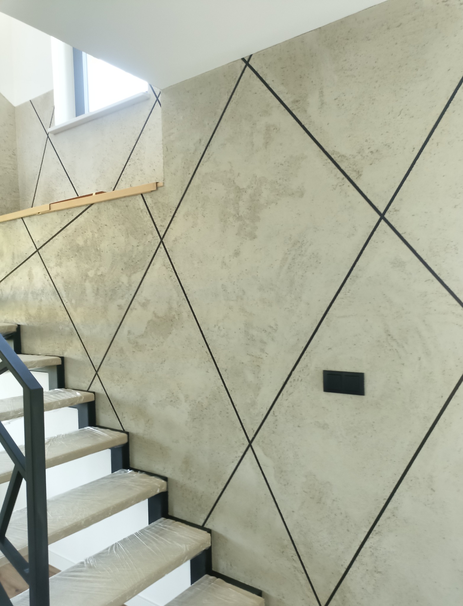 Ściana w korytarzu wykończona gładzią dekoracyjną w odcieniach szarości, z czarnymi liniami tworzącymi geometryczny wzór rombów, widoczne schody zabezpieczone folią ochronną.