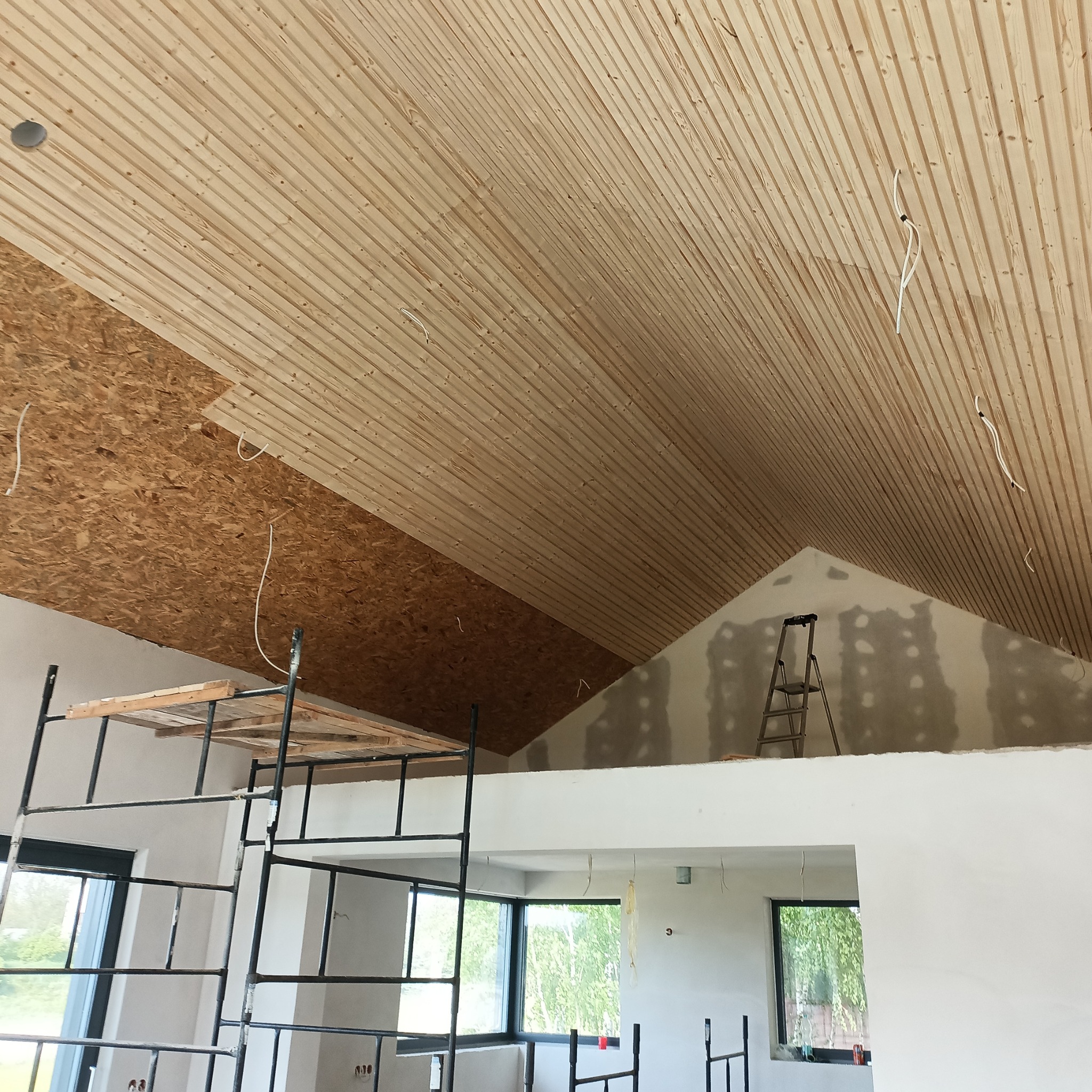 Skośny sufit z drewnianą boazerią w trakcie montażu, częściowo wykończony, widoczne kable, rusztowanie i drabina w surowym wnętrzu domu.