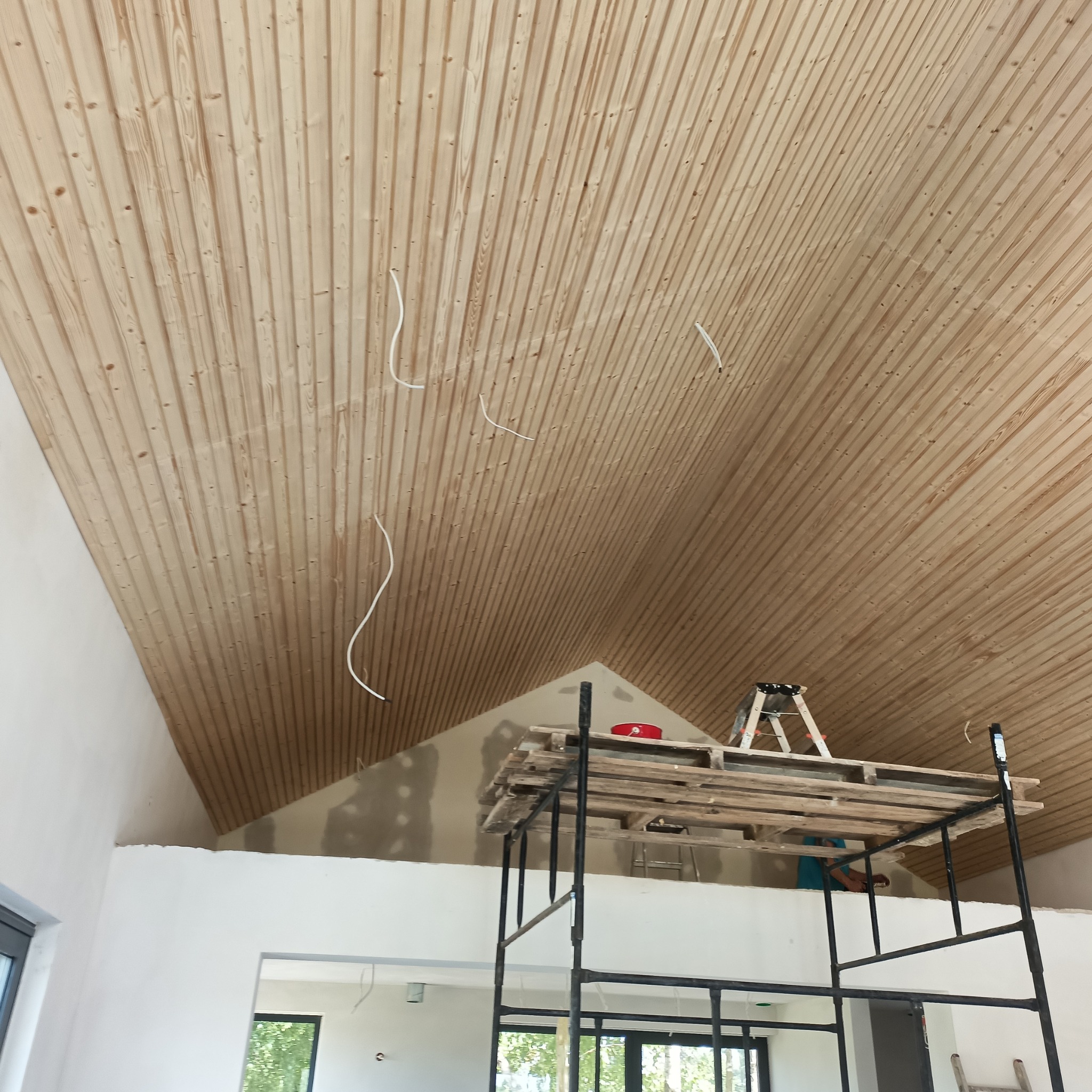 Drewniana boazeria na skośnym suficie poddasza, widoczne kable elektryczne, rusztowanie z palet i drabina w tle, ślady szpachlowania na ścianie.