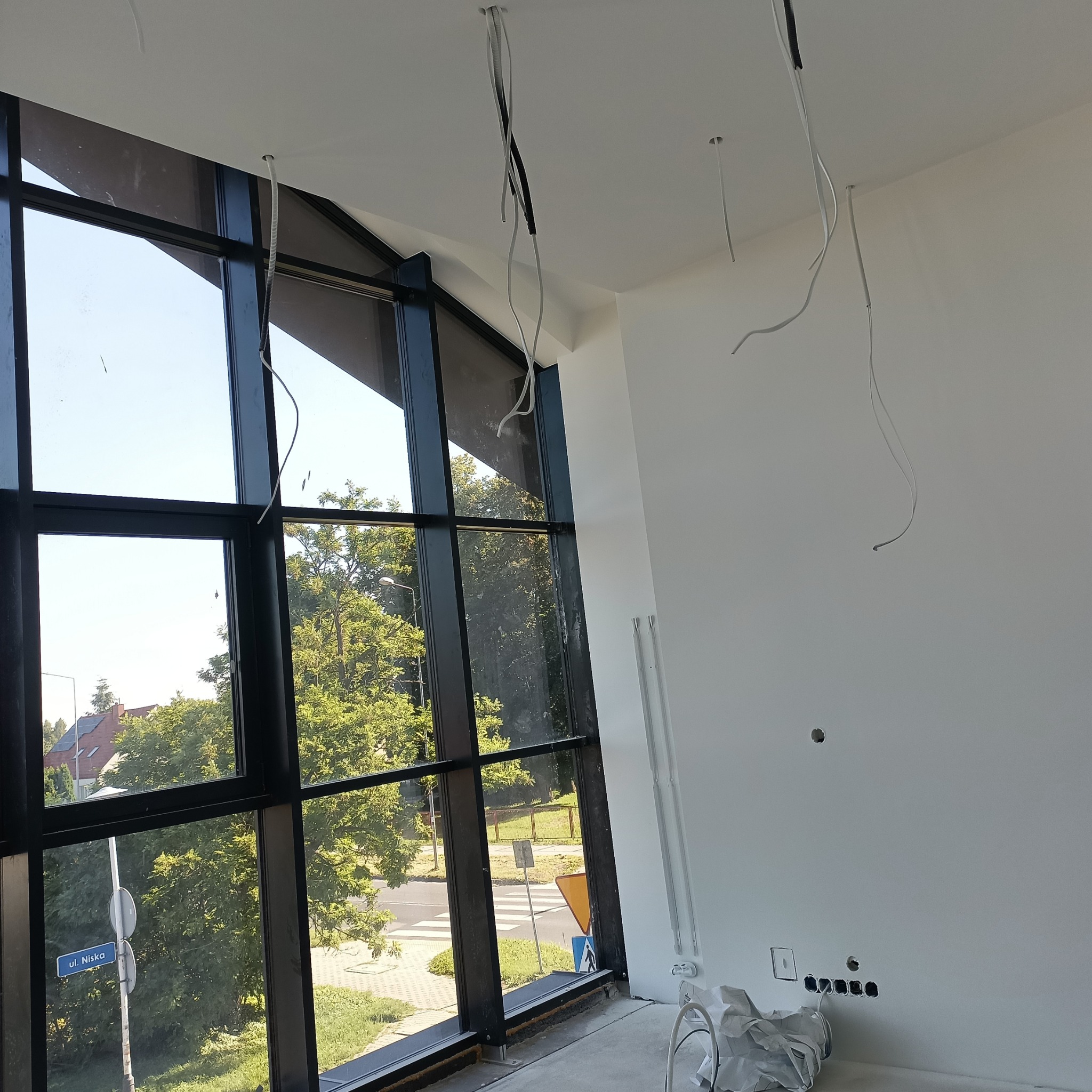 Wnętrze w trakcie wykończenia: świeżo pomalowane białe ściany, duże okno z czarną ramą z widokiem na ulicę i zieleń, wystające kable elektryczne z sufitu i ściany, przygotowane gniazdka elektryczne.