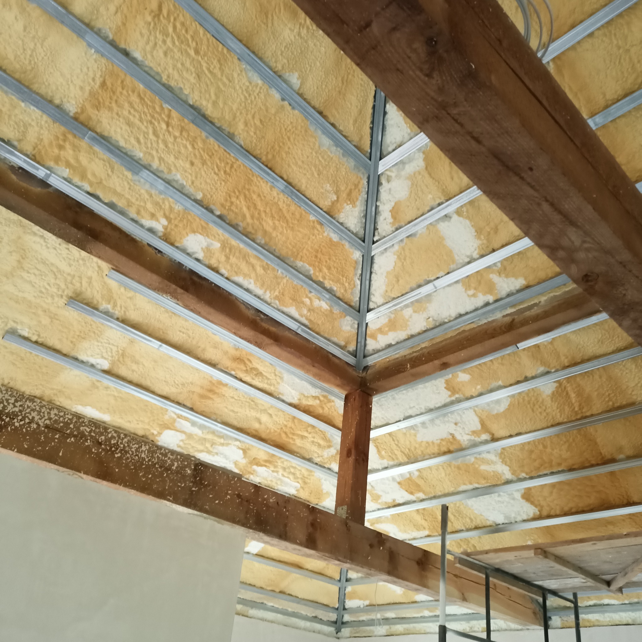 Poddasze z widoczną konstrukcją metalową i drewnianą, pokryte żółtą pianką izolacyjną z białymi plamami, widok od dołu.