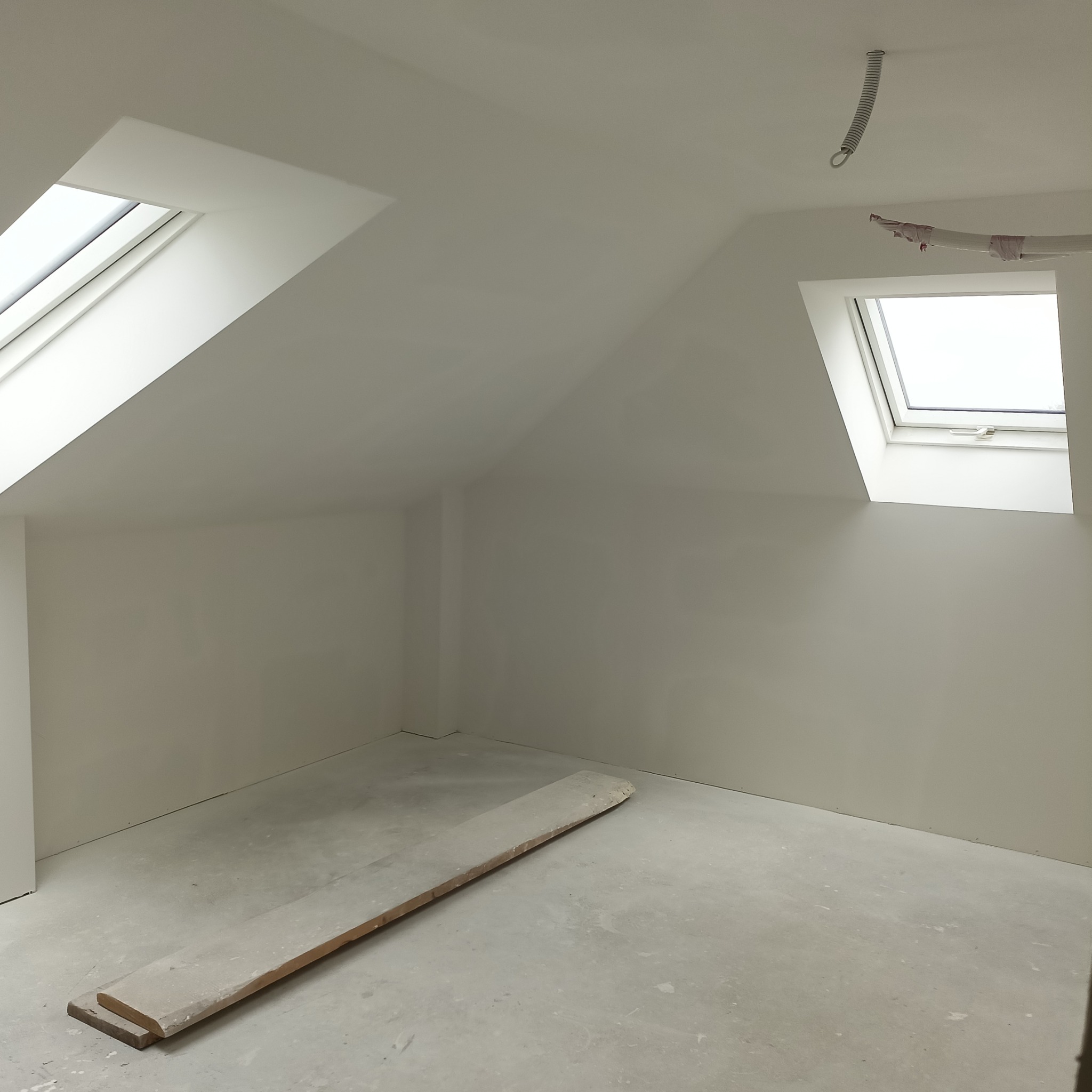 Poddasze po wykonaniu gładzi, widoczne okna dachowe, surowa podłoga i zwisający przewód oświetleniowy.