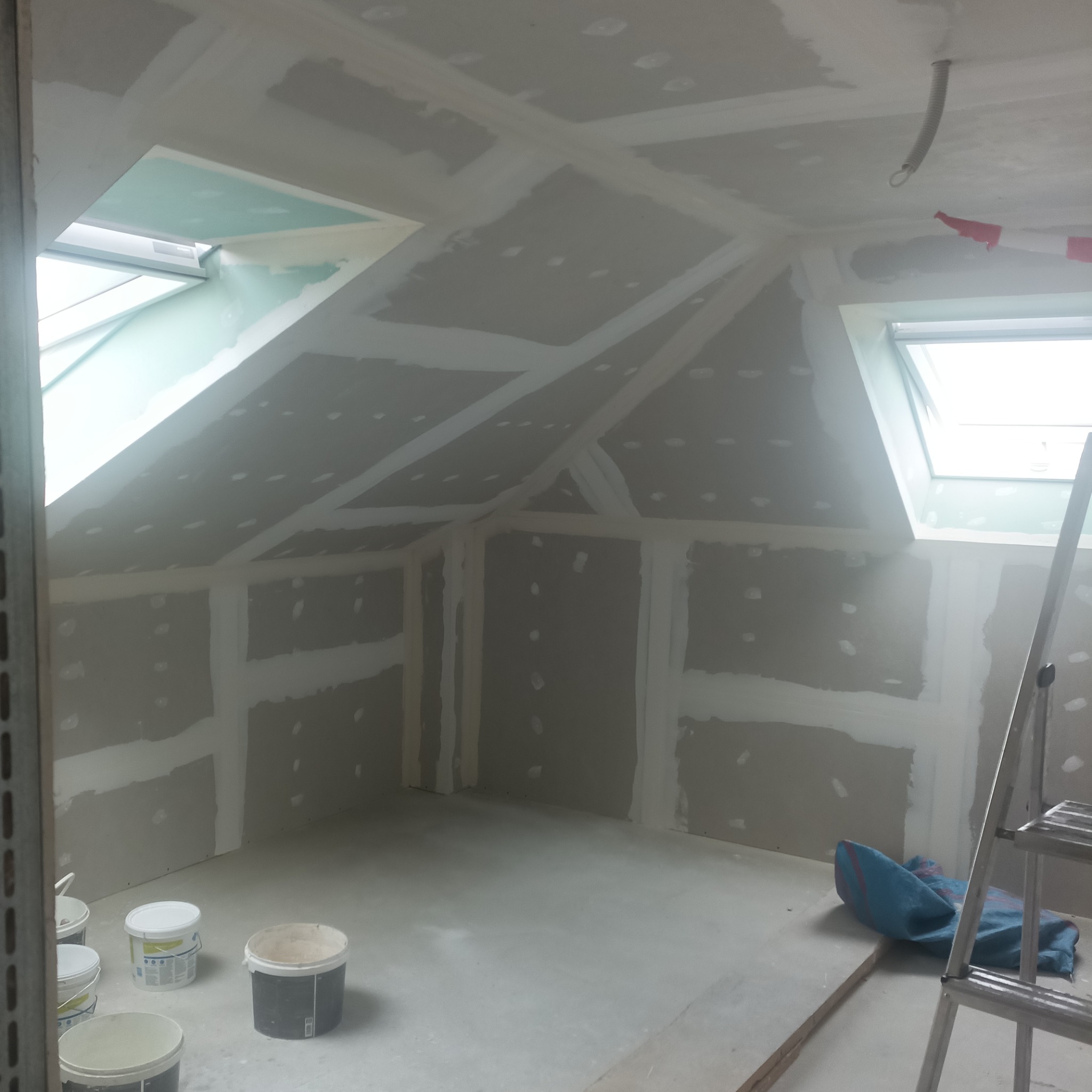 Poddasze w trakcie prac wykończeniowych: zamontowane płyty gipsowo-kartonowe, widoczne łączenia wzmocnione taśmą, okna dachowe, drabina i wiaderka z masą szpachlową.