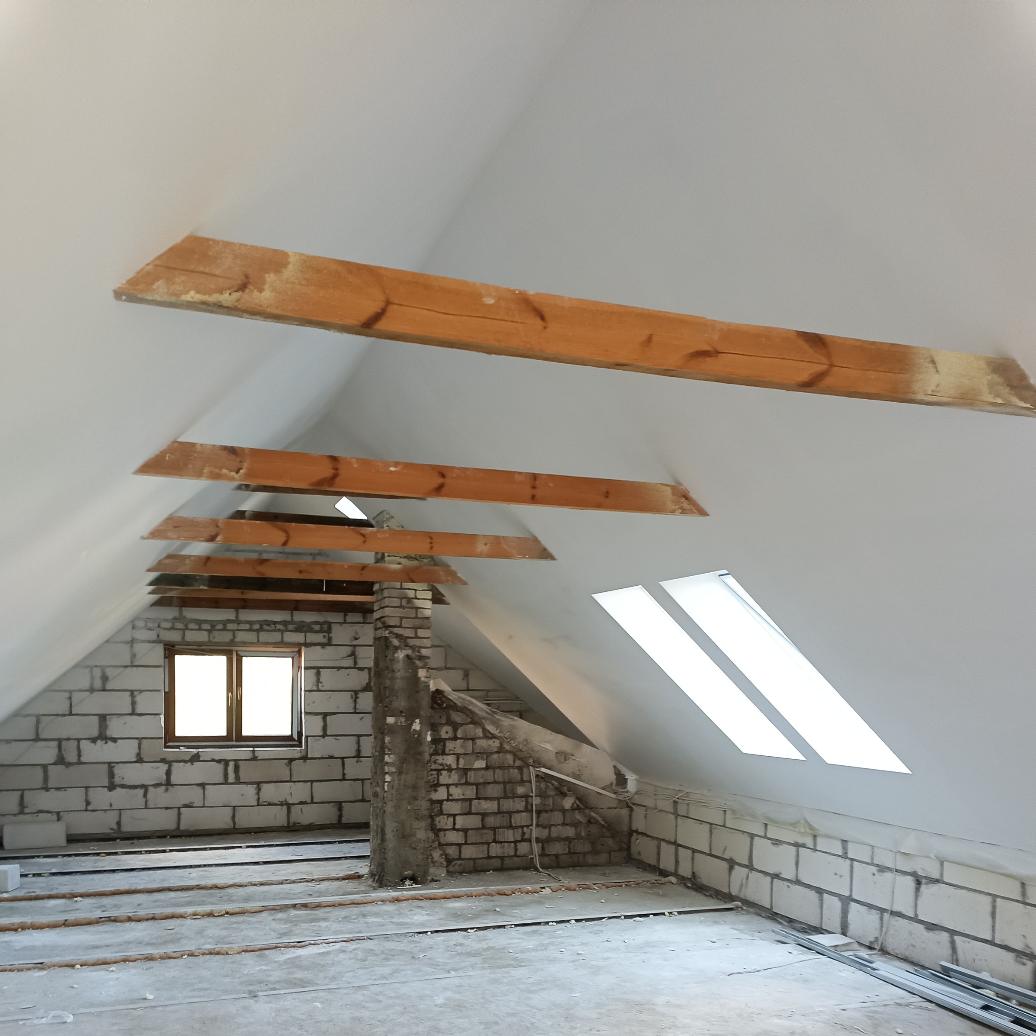 Poddasze w trakcie adaptacji: surowe ściany z bloczków, drewniane belki stropowe, okno dachowe i okno w ścianie szczytowej, widoczne ślady prac budowlanych.
