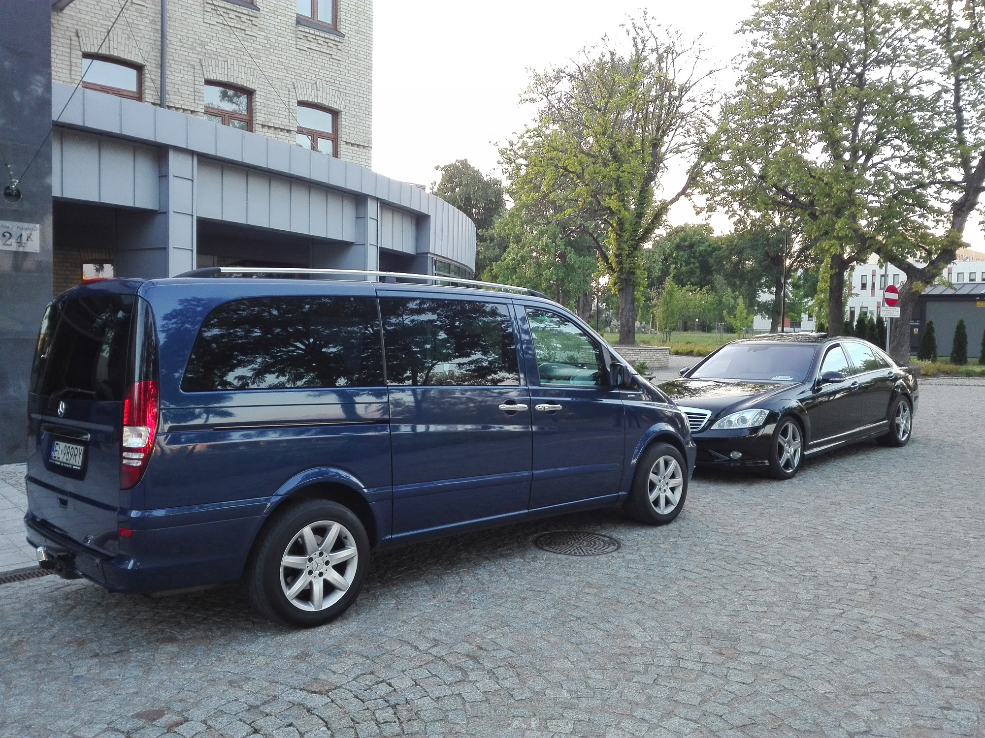 Granatowy van Mercedes-Benz Vito i czarna limuzyna Mercedes-Benz S-Class zaparkowane na brukowanej nawierzchni przed budynkiem.