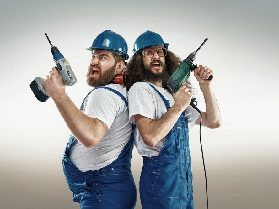Dwóch mężczyzn w niebieskich kombinezonach i kaskach, stojących tyłem do siebie, trzyma w rękach wiertarki i krzyczy.