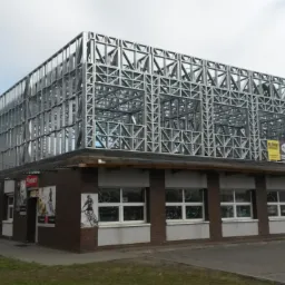 Stalowa konstrukcja nadbudowy na istniejącym budynku z oknami, widoczne elementy montażowe i niewielka reklama na górze konstrukcji.