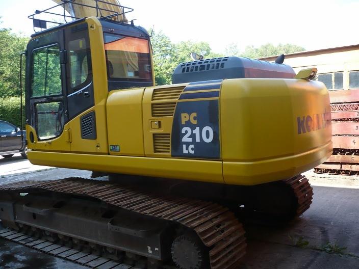 Żółta koparka gąsienicowa Komatsu PC210 LC-8, widok z boku na kabinę i tylną część maszyny, na tle drzew i budynku.