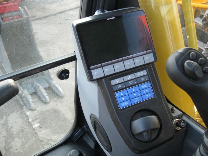 Wnętrze kabiny koparki gąsienicowej Komatsu PC210 LC-8, widoczny panel sterowania z ekranem, przyciski funkcyjne, joystick i fragment szyby z widocznym łyżką koparki na zewnątrz.