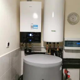 Zestaw urządzeń grzewczych: pompa ciepła Daikin z panelem sterowania, biały zasobnik ciepłej wody i szary zbiornik buforowy Galmet, zainstalowane w kotłowni z płytkami ceramicznymi na ścianach.