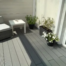 Szary taras z desek kompozytowych z wiklinowym fotelem z szarą poduszką, białym stolikiem kawowym z trzema szklankami, doniczkami z roślinami i kwiatami, przylegający do białej ściany i okna.