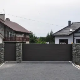 Automatyczna brama przesuwna z poziomymi panelami w kolorze grafitowym, osadzona w kamiennych murkach z łupka, prowadząca na posesję z domami w tle.