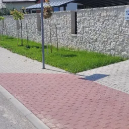 Nowo ułożona czerwona kostka brukowa na chodniku sąsiadująca z szarą kostką, tłem jest kamienny mur z numerem domu i młodymi drzewkami.