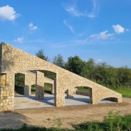 Kamienna konstrukcja z łukami w trakcie budowy na zewnątrz, widoczne elementy betonowe i ziemia wokół.
