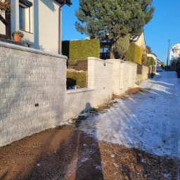 Nowo wybudowany, długi, niski murek z białej cegły wzdłuż drogi z resztkami śniegu, widok na domy i drzewa w tle.