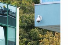 Zewnętrzna kamera obrotowa PTZ zamontowana na błękitnej ścianie budynku, widok z dołu, w tle zieleń drzew.