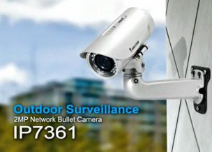 Zewnętrzna kamera cylindryczna IP7361, zamontowana na ścianie budynku, z widocznym obiektywem i uchwytem, na tle rozmytego budynku biurowego i błękitnego nieba.