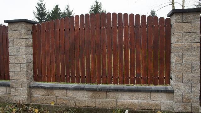 Drewniane ogrodzenie panelowe z brązowych desek, osadzone na kamiennej podmurówce i słupkach z szarego kamienia, na tle zieleni drzew w pochmurny dzień.
