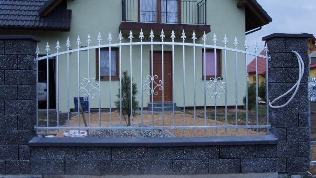 Metalowe ogrodzenie z dekoracyjnymi elementami i słupkami z szarej kostki, na tle jasnego domu z brązowymi oknami i drzwiami.