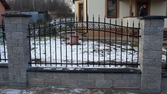 Metalowe ogrodzenie z dekoracyjnymi elementami i kamiennymi słupkami. Częściowo ośnieżony teren. W tle dom jednorodzinny i drzewa.