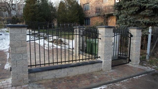 Nowoczesne ogrodzenie z czarnymi, metalowymi przęsłami i słupkami z szarej kostki łupanej, zwieńczonymi czarnymi daszkami. Brama z dekoracyjnymi elementami.