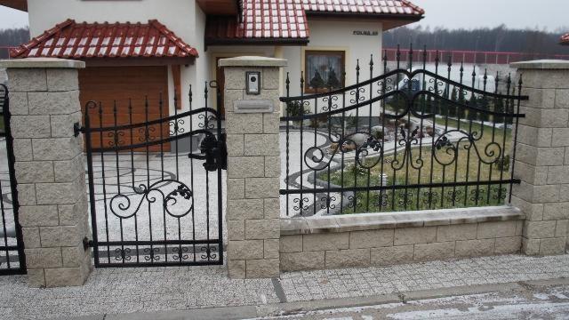 Kuta, czarna brama z dekoracyjnym motywem roślinnym i kamienne słupki na tle domu z czerwoną dachówką i szarym chodnikiem. Zimowa sceneria.