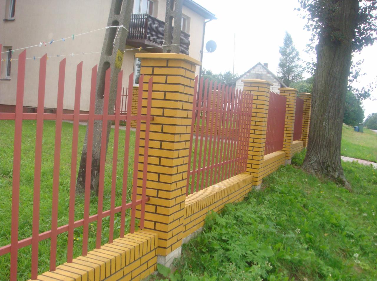 Żółto-ceglany murek z czerwonym, metalowym ogrodzeniem palisadowym, widok z perspektywy trawnika. Słupki ogrodzenia z cegły, w tle dom.