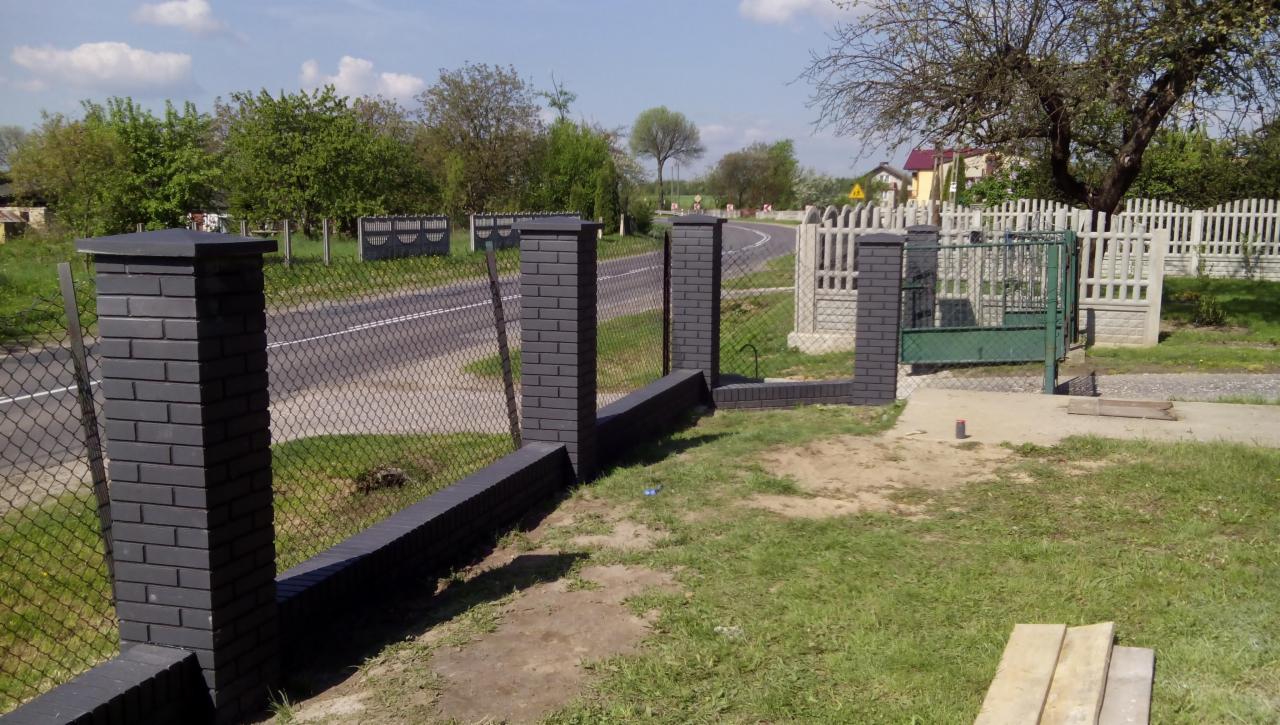Nowoczesne ogrodzenie z czarnych cegieł klinkierowych i siatki, z bramą w tle. Słupki ogrodzeniowe z ciemnej cegły. Widok na drogę i zieleń.