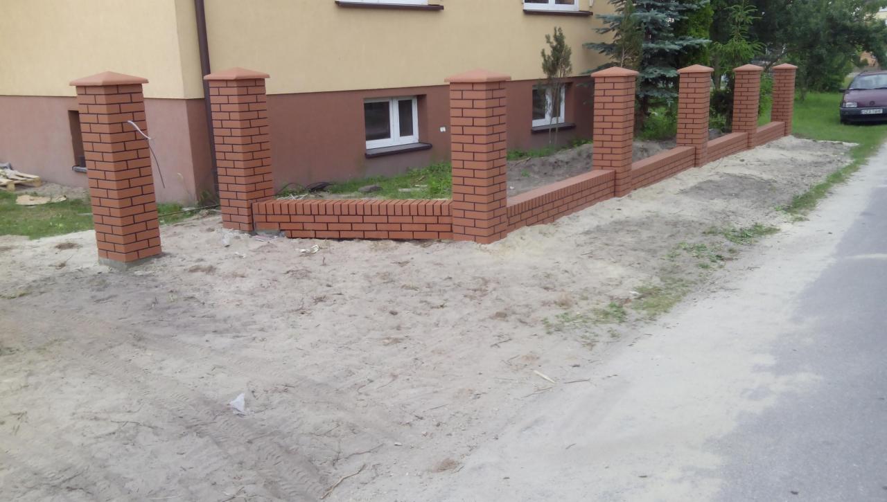 Ceglany, częściowo ukończony murek ogrodzeniowy z filarami, widoczny z perspektywy ulicy, z budynkiem w tle. Elementy murku w kolorze czerwonej cegły.
