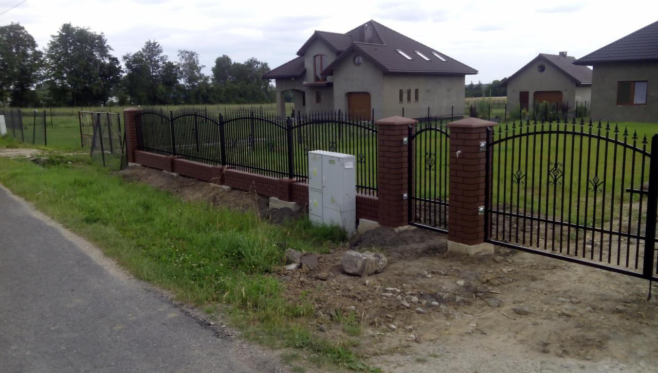 Nowoczesne ogrodzenie z czarnymi przęsłami i ceglanymi słupkami, brama wjazdowa. W tle domy jednorodzinne w stanie surowym otwarte.