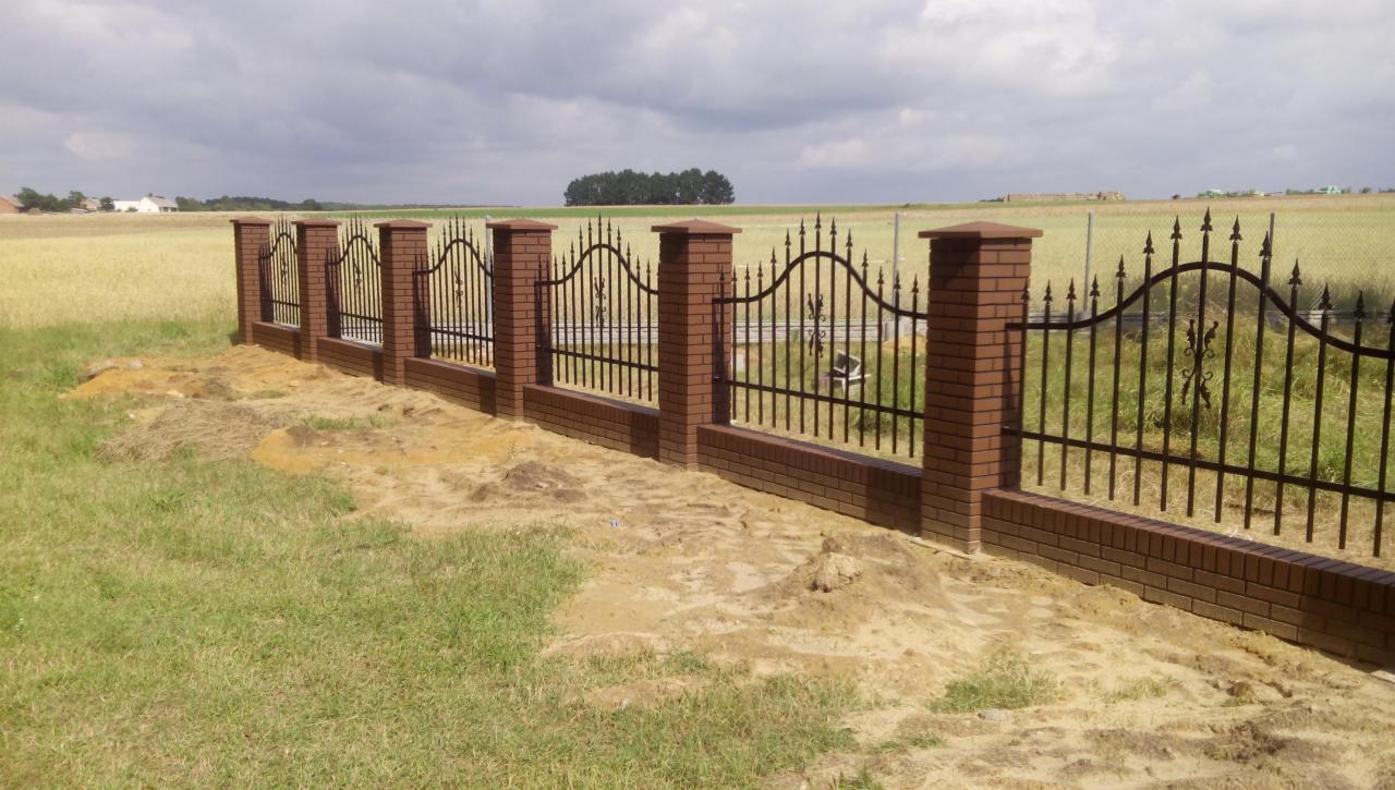 Nowoczesne ogrodzenie z cegły klinkierowej i metaloplastyki na tle zielonego pola. Elementy dekoracyjne i solidna konstrukcja.