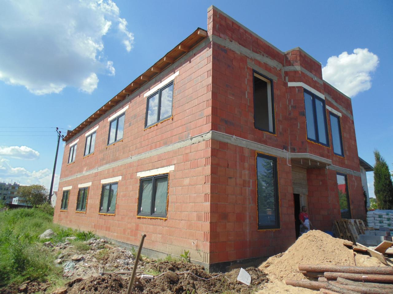 Dwukondygnacyjny dom w trakcie budowy z czerwonej cegły, z zamontowanymi oknami z czarną ramą. Widoczne materiały budowlane i częściowo ułożone drewno.