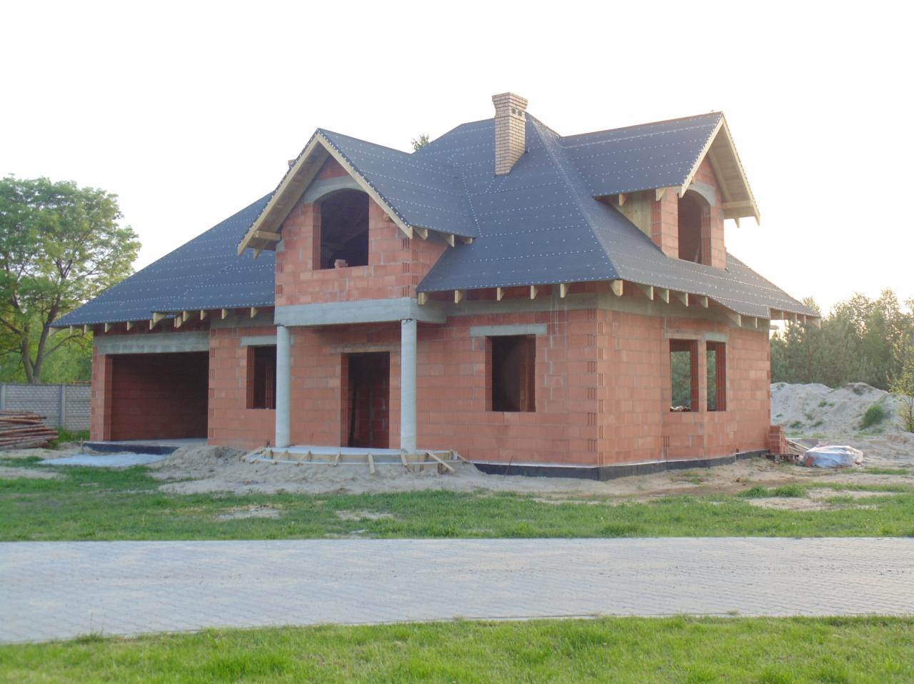 Dom murowany w trakcie budowy: czerwona cegła, czarny dach, niezadaszony garaż, widoczne elementy konstrukcyjne, trawa i kostka brukowa przed budynkiem.