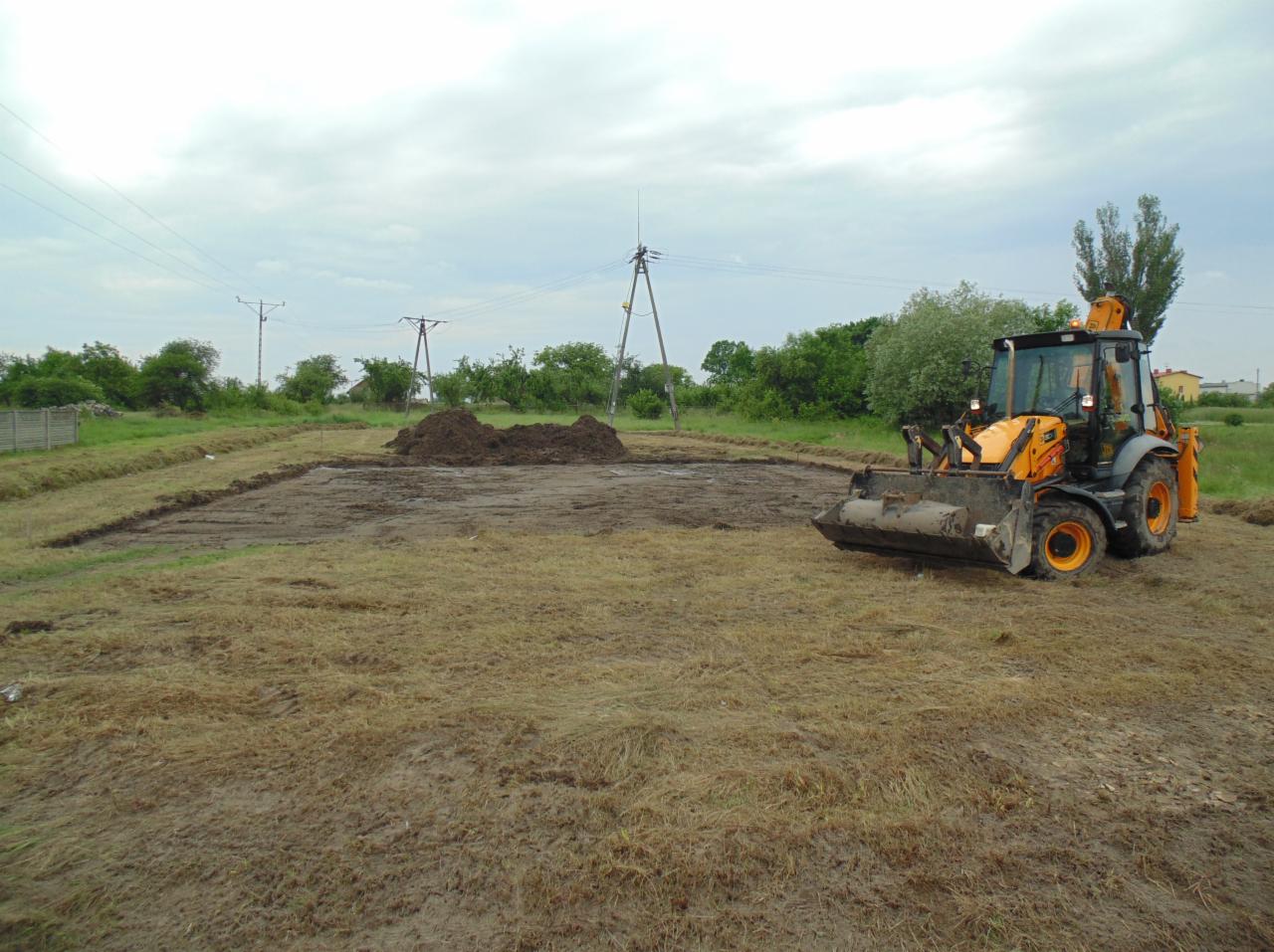 Żółta koparko-ładowarka na przygotowanym terenie pod budowę, z wyznaczonymi granicami i stertą ziemi w tle. Widoczne słupy energetyczne.